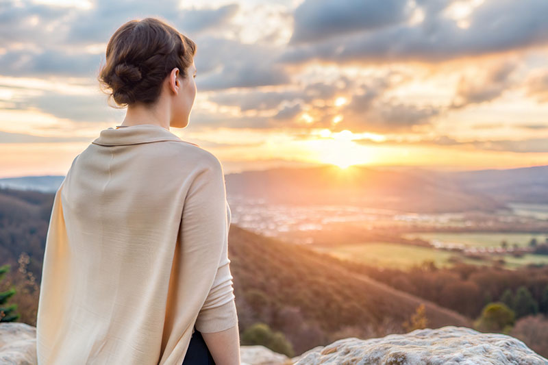 Eine Frau steht auf einem Felsen und blickt über ein weites Tal in Richtung Sonnenuntergang.