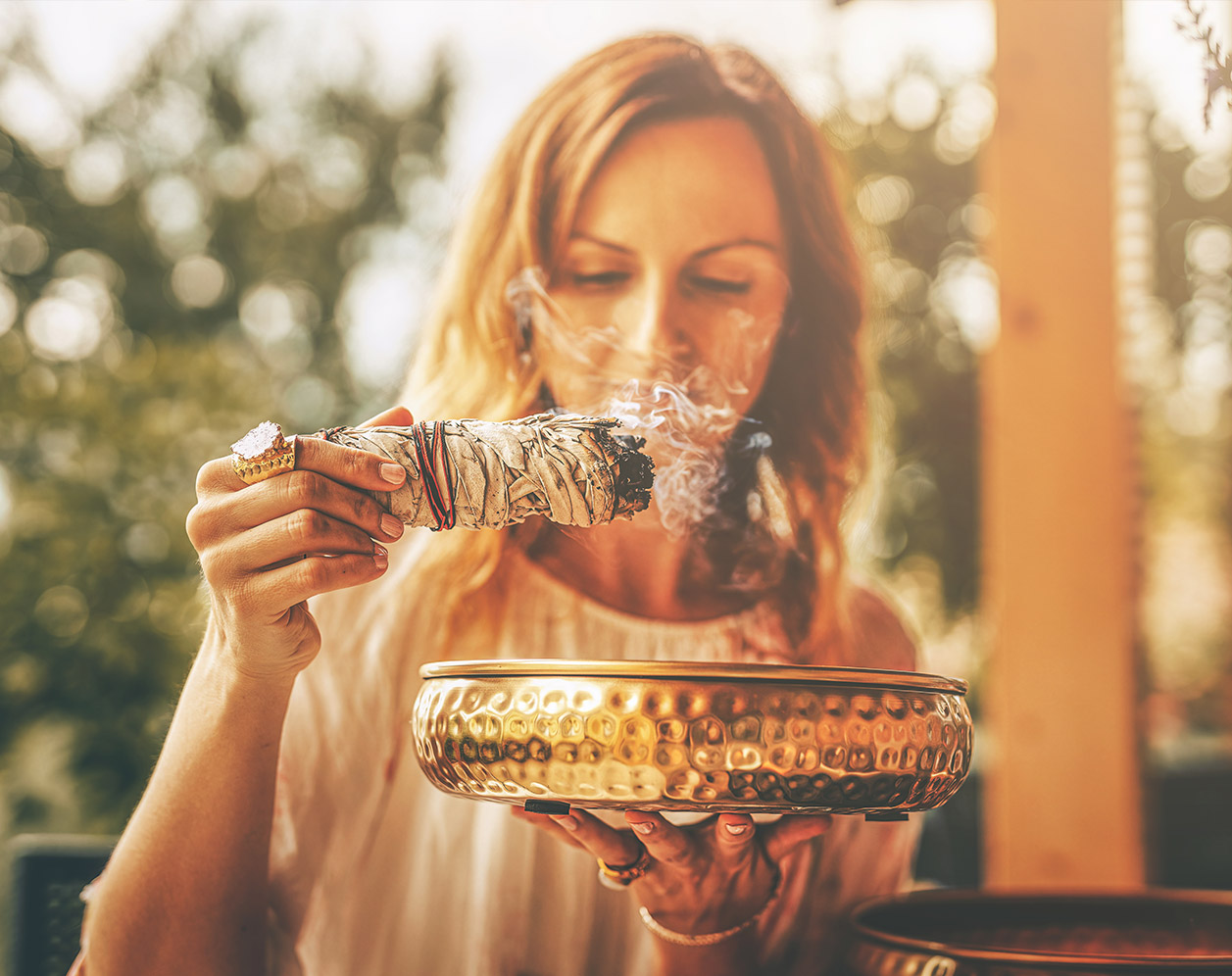 Eine Frau hält eine brennende Salbeibündel in der Hand, während sie eine goldene Schale darunter hält. Rauch steigt sanft auf, während das Licht einer sonnigen Umgebung die Szene durchflutet.